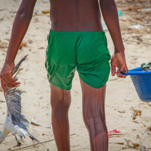 Photographie de Madagascar. Village de pêcheurs de Morondava. Adolescent de dos, avec seulement un short vert mouillé. Il tient dans sa main gauche une mouette morte, et dans la droite l'une des anses d'un bassine pleine de poussons.