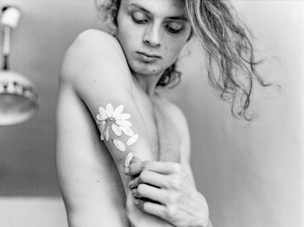 Photographie d'un jeune homme torse nu, pétals de marguerite collés sur son bras. Il tente de les décoller.