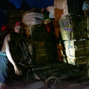Photographie de Madagascar. Diego-Suarez la nuit. Des hommes poussent un chariot très chargé, éclairés par les phares d'une voiture.