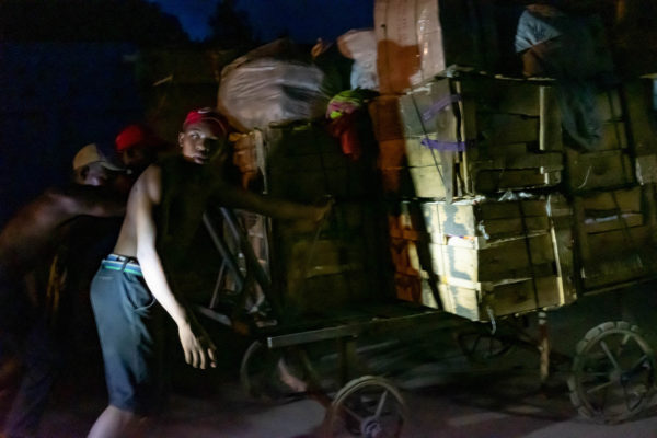 Photographie de Madagascar. Diego-Suarez la nuit. Des hommes poussent un chariot très chargé, éclairés par les phares d'une voiture.