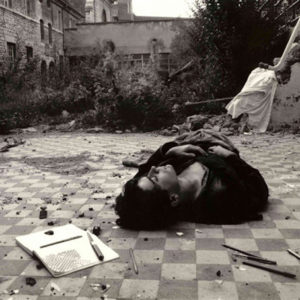 Photographie argentique noir et blanc.Un jeune homme habillé de noir est allongé endormi sur un carrelage en damiers dans une pièce à ciel ouvert. Il a reproduit sur un cahier petit careaux le damier du sol.