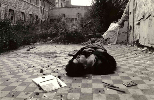 Photographie argentique noir et blanc.Un jeune homme habillé de noir est allongé endormi sur un carrelage en damiers dans une pièce à ciel ouvert. Il a reproduit sur un cahier petit careaux le damier du sol.