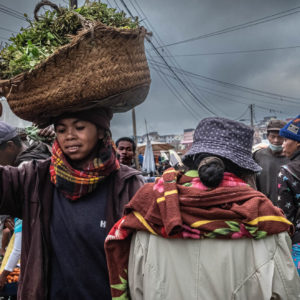 Madagascar, le marché des paysans. Au premier plan, une jeune fille souriante porte sur sa tête un grand panier rempli de brèdes.