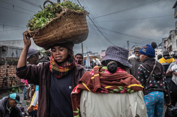 Madagascar, le marché des paysans. Au premier plan, une jeune fille souriante porte sur sa tête un grand panier rempli de brèdes.
