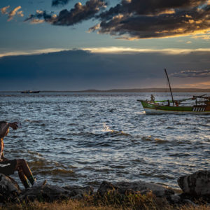 Un jeune homme tend le bras pour faire un selfie en bord de mer, au soleil couchant. Sur l'eau, un boutre sur lequel les marins s'activent.