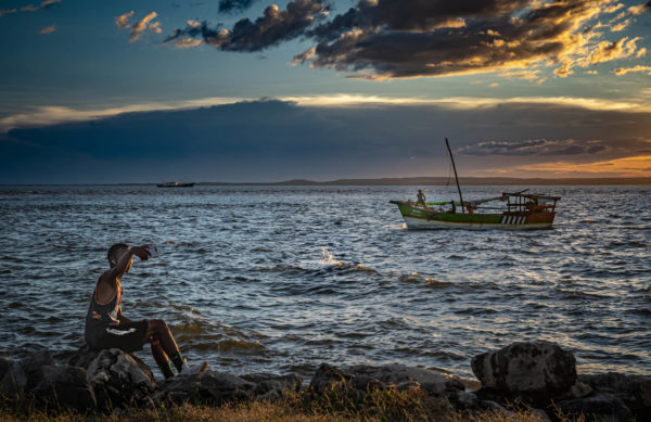 Un jeune homme tend le bras pour faire un selfie en bord de mer, au soleil couchant. Sur l'eau, un boutre sur lequel les marins s'activent.